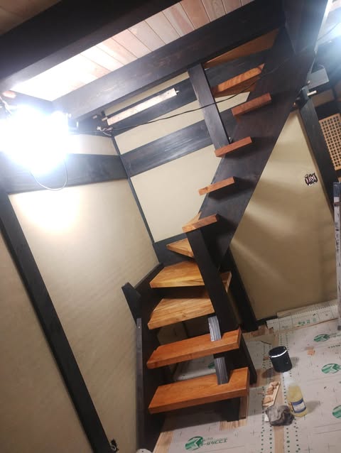 Black metal staircase with wooden steps rising at an angle between tan walls in a partially finished space; construction materials and exposed beams visible on the floor.