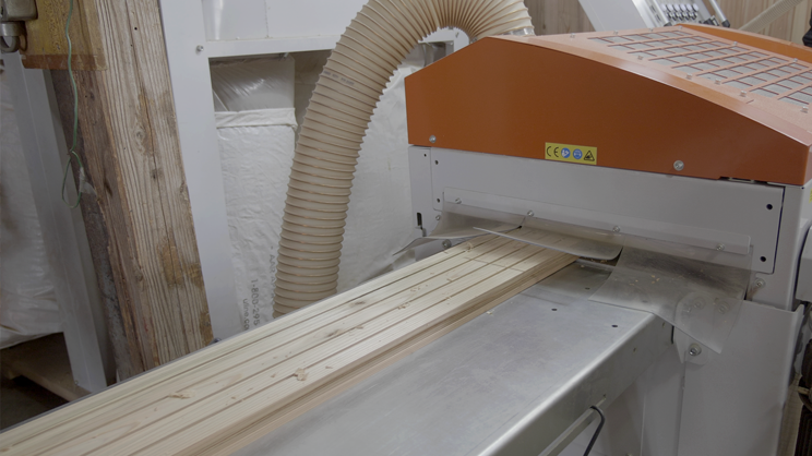 image - NO1製材機ウッドマイザー正規販売代理店│ウッドマイザージャパン MSco.,LTD. Wood boards are fed into an industrial wood planer in a workshop, with an orange-top machine guiding the boards downstream.