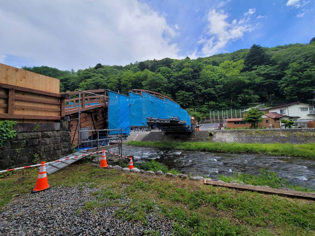 清流の上に架かる修復中の木造アーチ橋と青空・長野の山々の風景